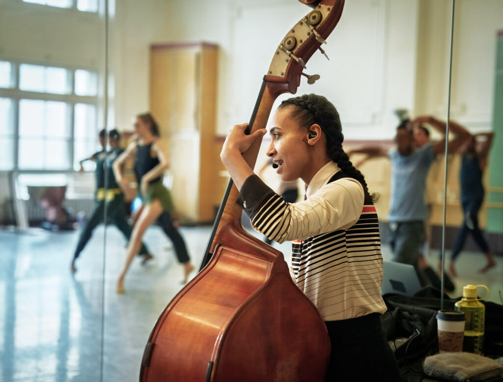 esperanza spalding plays her bass and sings into a microphone while watching dancers rehearse. In the mirror reflection behind her, the dancers step forward onto a bent leg in forced arch, their arms creeping upward.