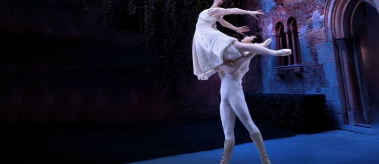 In an Italian-style courtyard, two dancers perform a pas de deux. Zecheng Liang lifts Thays Golz overhead as she tucks one leg into retiré and extends the other back in arabesque. She arches up and back with her arms wide behind her. Both dancers wear white-colored costumes and look up.