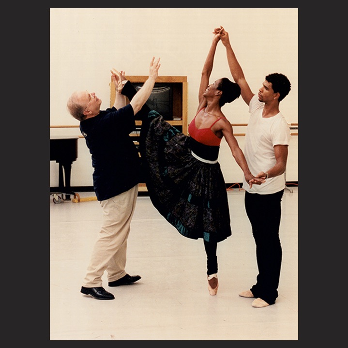 Ben Stevenson, in a black polo shirt and khakis, adjusts the foot of ballerina Lauren Anderson as she and Carlos Acosta, both in practice clothes, practice a pas de deux.