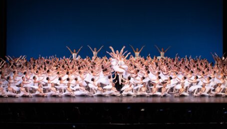 A large group of young ballet students wearing white tutus poses in a huge tableau. A line of girls in the center are being lifted up by the waist by partners in black tights.