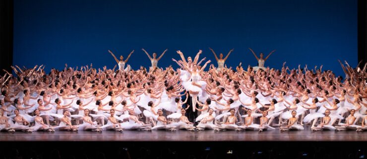 A large group of young ballet students wearing white tutus poses in a huge tableau. A line of girls in the center are being lifted up by the waist by partners in black tights.