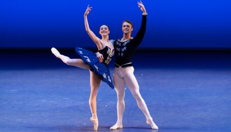 Two dancers perform a pas de deux onstage. A woman in a royal blue tutu does an attitude derrière on pointe in profile as her partner, who wears white tights and ballet shoes and a dark blue tunic, holds her around the waist as he stands in tednu.