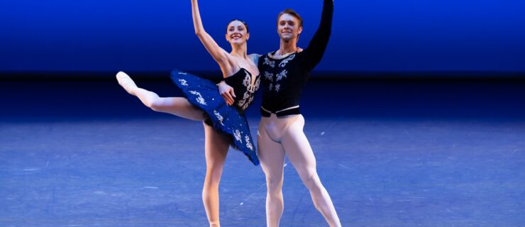 Two dancers perform a pas de deux onstage. A woman in a royal blue tutu does an attitude derrière on pointe in profile as her partner, who wears white tights and ballet shoes and a dark blue tunic, holds her around the waist as he stands in tednu.