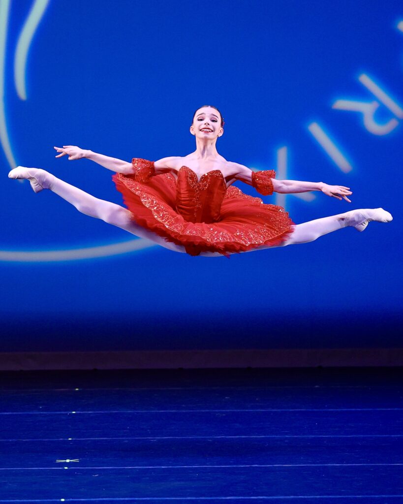 Against a blue background on a stage, a young female dancer performs a jump in second position, her legs extended past 180 degrees and her arms in line with her legs. She wears a red tutu.