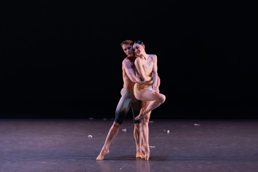 A ballerina in a ruched, beiige leotard poses in passé parallel on pointe as her partner, wearing gray, knee-length tights, stands behind her in tendu and wraps his arms around her.