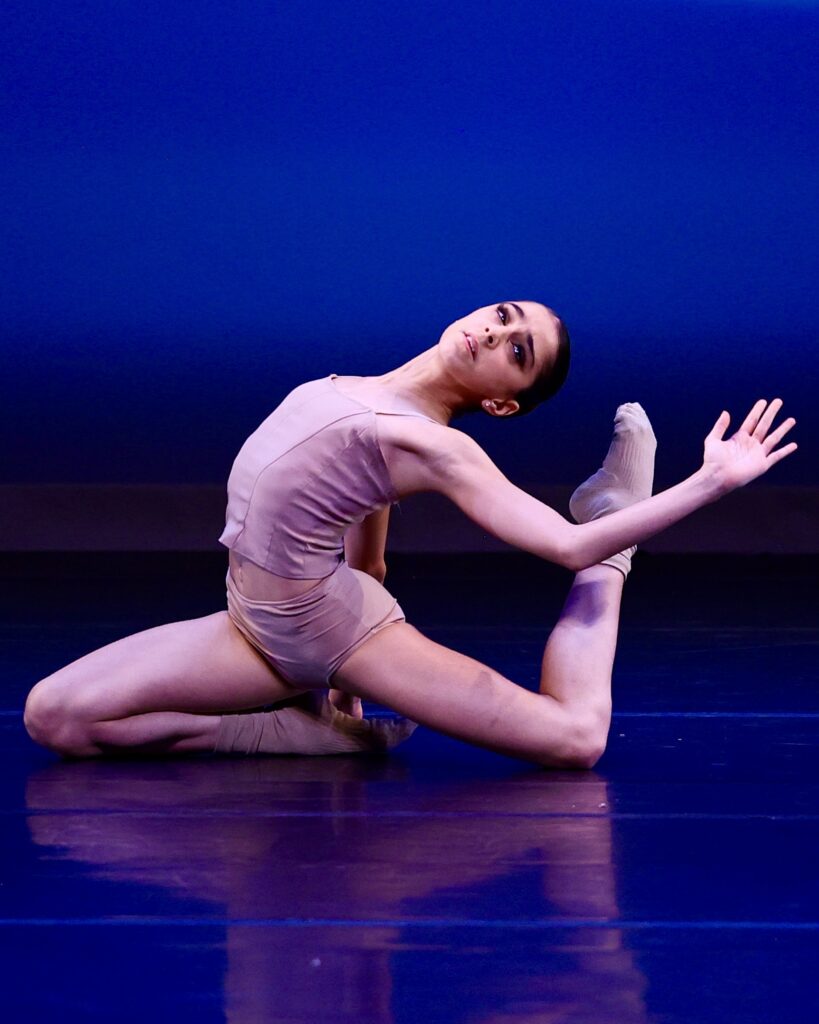 A dancer kneels on her front knee on a stage, her back leg bent with her foot close to her head. She arches back towards her foot, with her left hand bent, palm facing the audience. She wears gray shorts, a gray tank, and gray ankle socks.