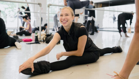 Calley Skalnik grins, leaning forward to rest one hand on her front foot as she sits in a split wearing ballet warmups.
