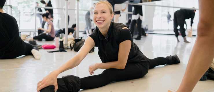 Calley Skalnik grins, leaning forward to rest one hand on her front foot as she sits in a split wearing ballet warmups.