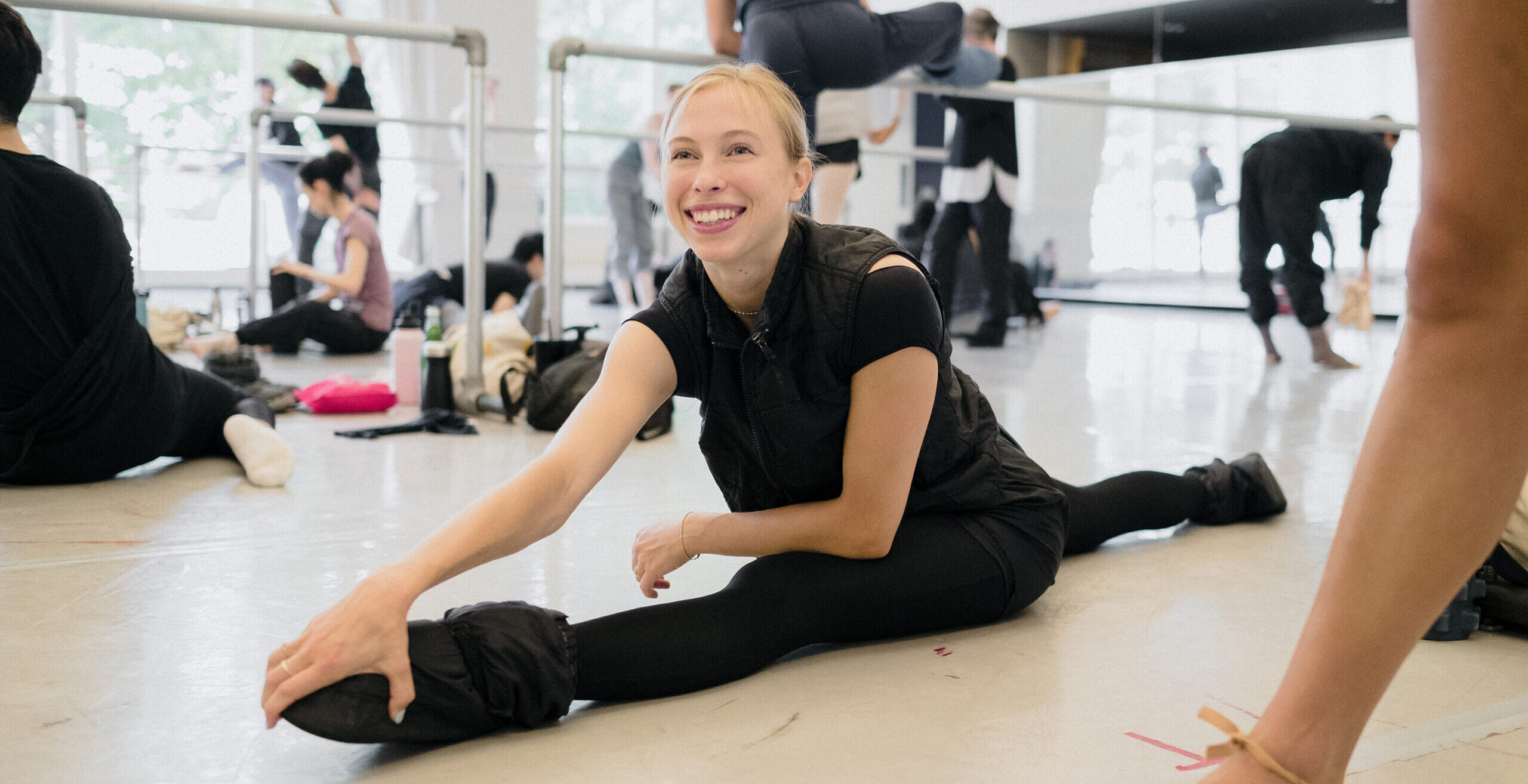 Calley Skalnik grins, leaning forward to rest one hand on her front foot as she sits in a split wearing ballet warmups.