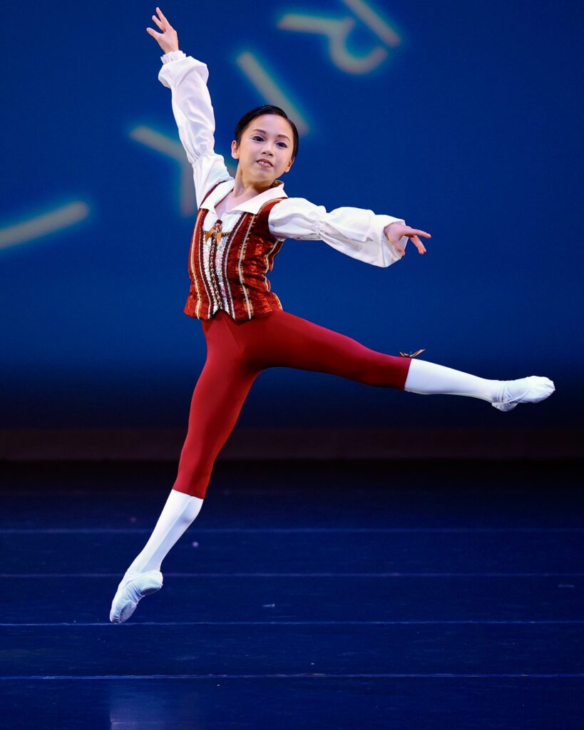 A young male dancer in red tights, white socks and shoes, and a red and white tunic performs a saute arabesque. He looks out at the audience.