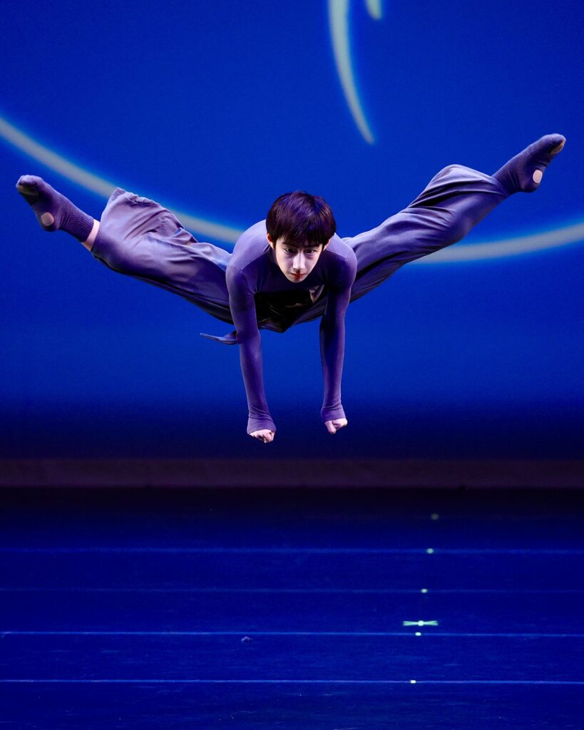 A young male dancer performs a jump in second position, his legs extended past 180 degrees and his arms punched down towards the floor, hands in fists. He wears loose blue/purple long pants and a matching long sleeve top.