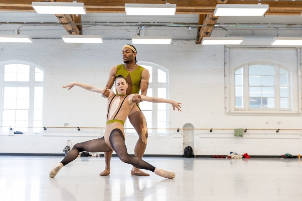 Vincent Van Harris and Grace Ann Powers dance a contemporary ballet pas de deux in a large dance studio with white walls. Van Harris holds Powers under her arms as she opens both legs to a front and back attitude, her pointed feet against the floor.