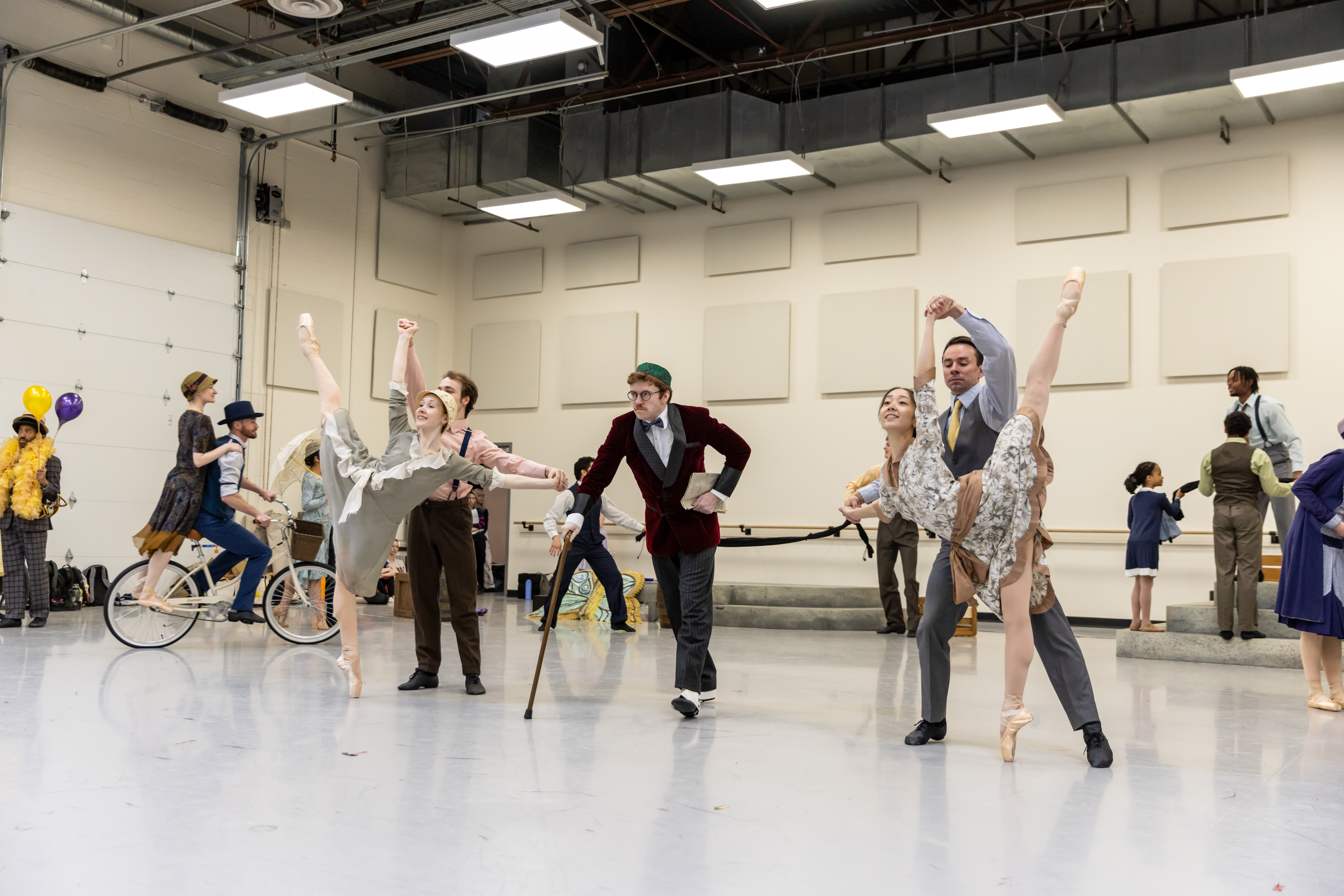 In a large ballet studio, two pairs of partners mirror each other doing supported arabesque penchés toward the center. Another dancer walks forward with a cane.