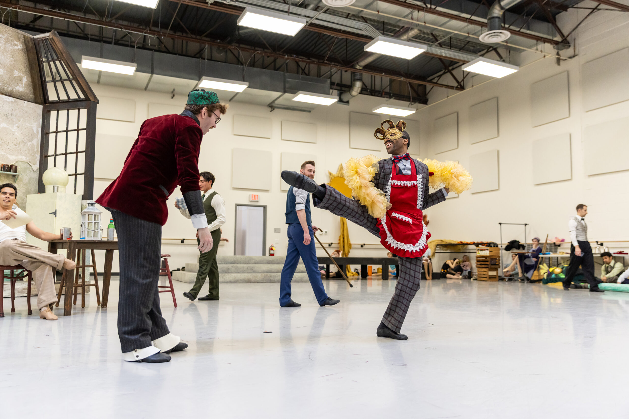 Two dancers—one wearing a red velvet blazer and a green hat, and the other with a red apron over a gray suit, a yellow boa, and a gold Mardi Gras mask—dance facing each other in a ballet studio.