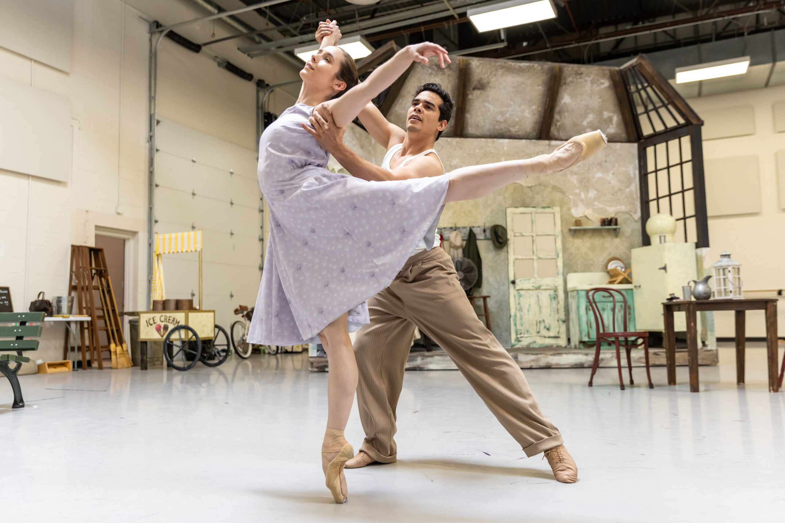 In a large ballet studio, Miguel Anaya partners Sophie Miklosovic as she does a piqué arabesque. She wears a light purple dress, and he tan slacks with a white tank top.