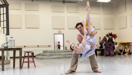 In a large ballet studio, Miguel Anaya holds Sophie Miklosovic in a dramatic fish pose. They both grin, she wearing a flowing light purple dress, and he tan slacks with a white tank top.