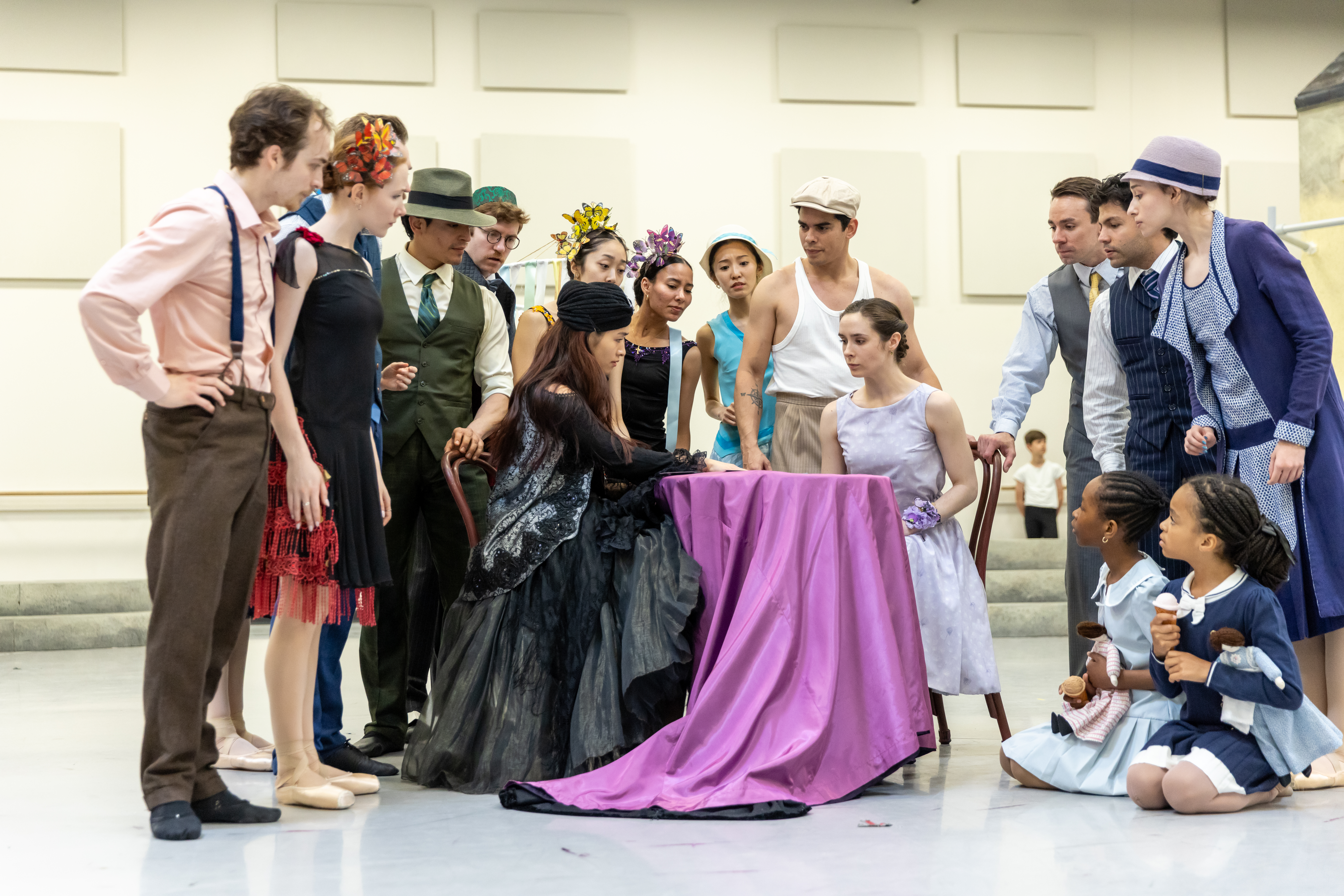 A group of ballet dancers dressed as 1920s townspeople gather around a small circular table with a bright purple tablecloth. On one side sits a fortuneteller, wearing a large and ornate black dress with lace. On the other is a woman in a light purple dress, who looks concerned by her fortune.