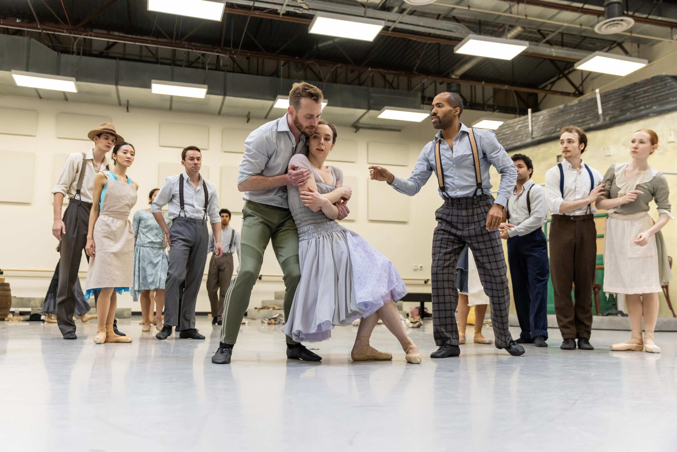 A group of dancers dressed in 1920s-style clothing gather around as one woman, in a light purple dress, falls back into her lover. She is ill and faint.