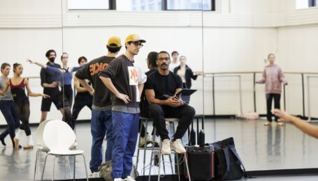Craig Hall sits on a stool in the front of a dance studio, holding an iPad and looking at the dancers in front of him. Justin Peck stands on his right side with his hands on his hips. The dancers, visible in the mirror, dance a sequence.