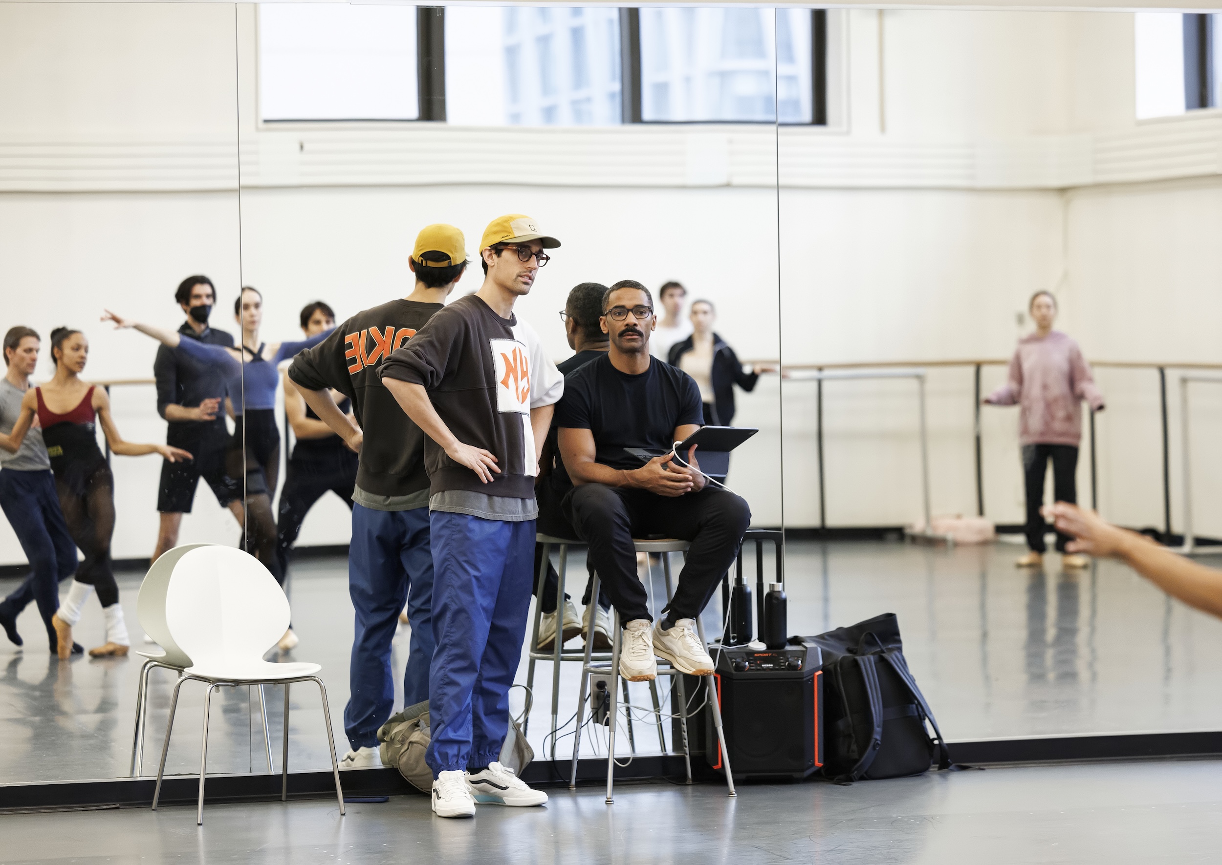 Craig Hall sits on a stool in the front of a dance studio, holding an iPad and looking at the dancers in front of him. Justin Peck stands on his right side with his hands on his hips. The dancers, visible in the mirror, dance a sequence.