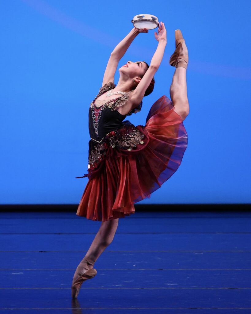 A young female dancer arches back in a dramatic back attitude, her front foot on pointe and her back foot nearly hitting the tambourine in her hands. She wears a red and black costume.