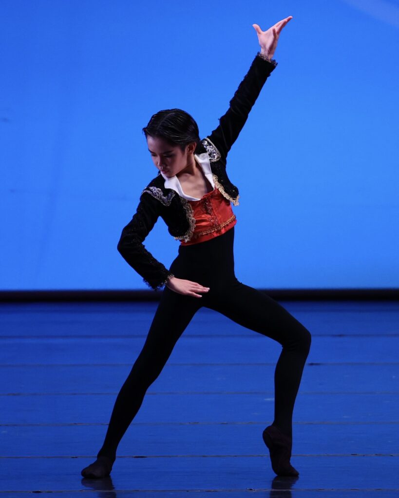 A young male dancer in black tights, a black bolero jacket and a red shirt stands in a wide fourth position, his front leg bent and heel up and his back leg straight. His front arm is up in a high V and his back arm is on his hip. He angles his head to the back arm.
