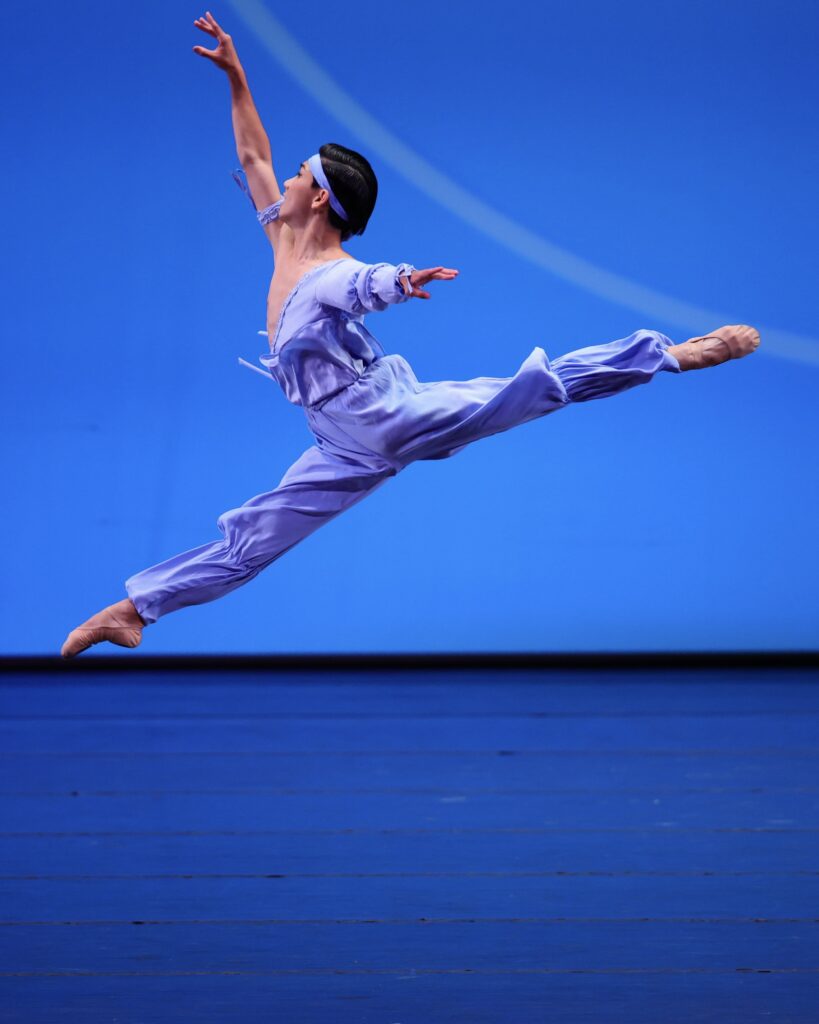 Against a blue backdrop on a stage, a boy dressed in loose blue pants, a one-shoulder blue shirt, and blue headband performs a saute arabesque