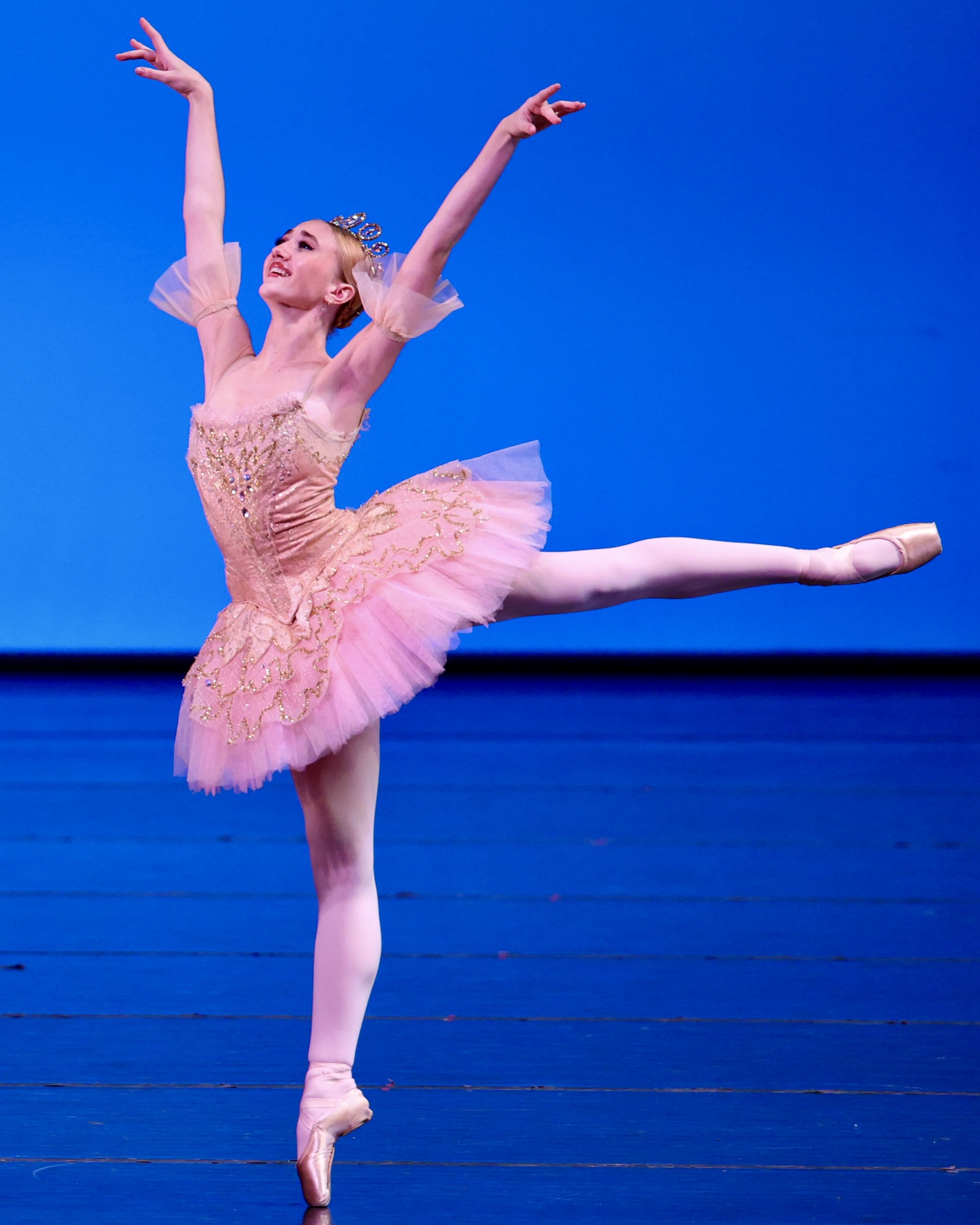 Against a blue background on a stage, a female dancer stands in arabesque on pointe, with her arms in a high V position and her gaze upwards. She wears a pink tutu.