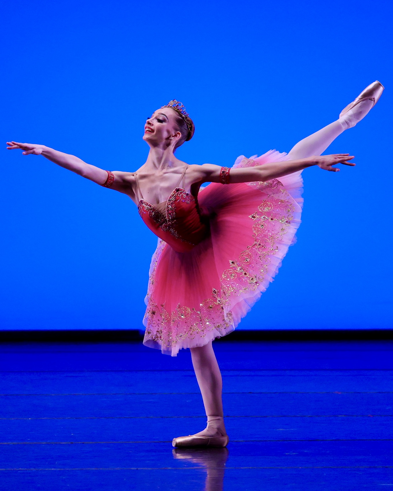 Against a blue background, a young female dancer in pointe shoes and a pink tutu plies in arabesque.