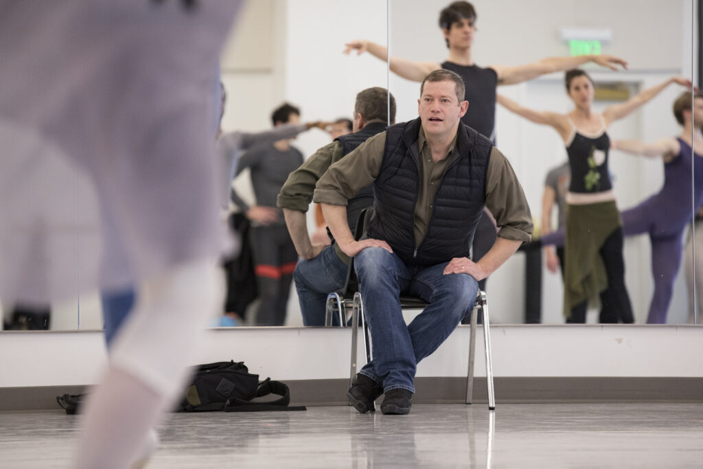Adam Sklute sits on a chair at the front of a ballet studio, his back to the mirror, as dancers move in front of him. He rests his hands on his thighs and leans slightly, his mouth open as he addresses them.