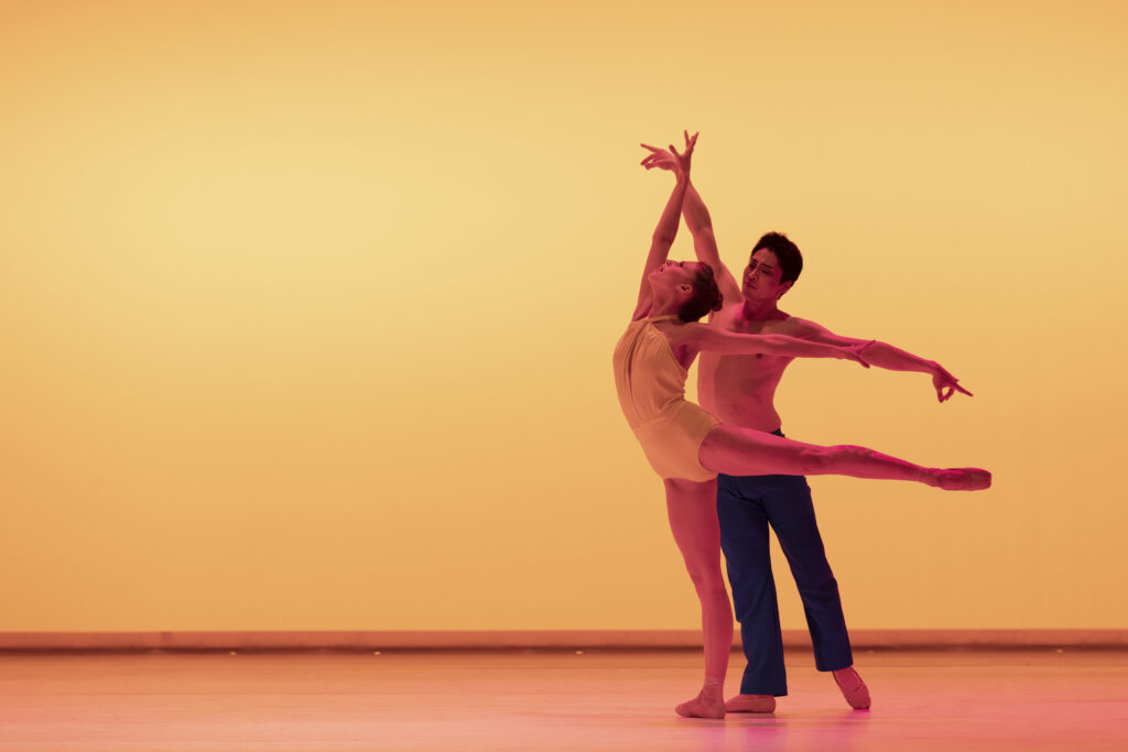 Onstage in front of a bright yellow backdrop, two dancers perform a contemporary pas de deux. Allison Perhach stands on flat with one leg in arabesque, looking up toward her right arm, which reaches toward the ceiling as her left arm reaches back. Behind her, Yoshiya Sakurai stands in tendu derriere with his arms in a wide third, pointer fingers extended gently. Perhach wears a yellow halter-top leotard, and Sakurai wears dark blue pants.