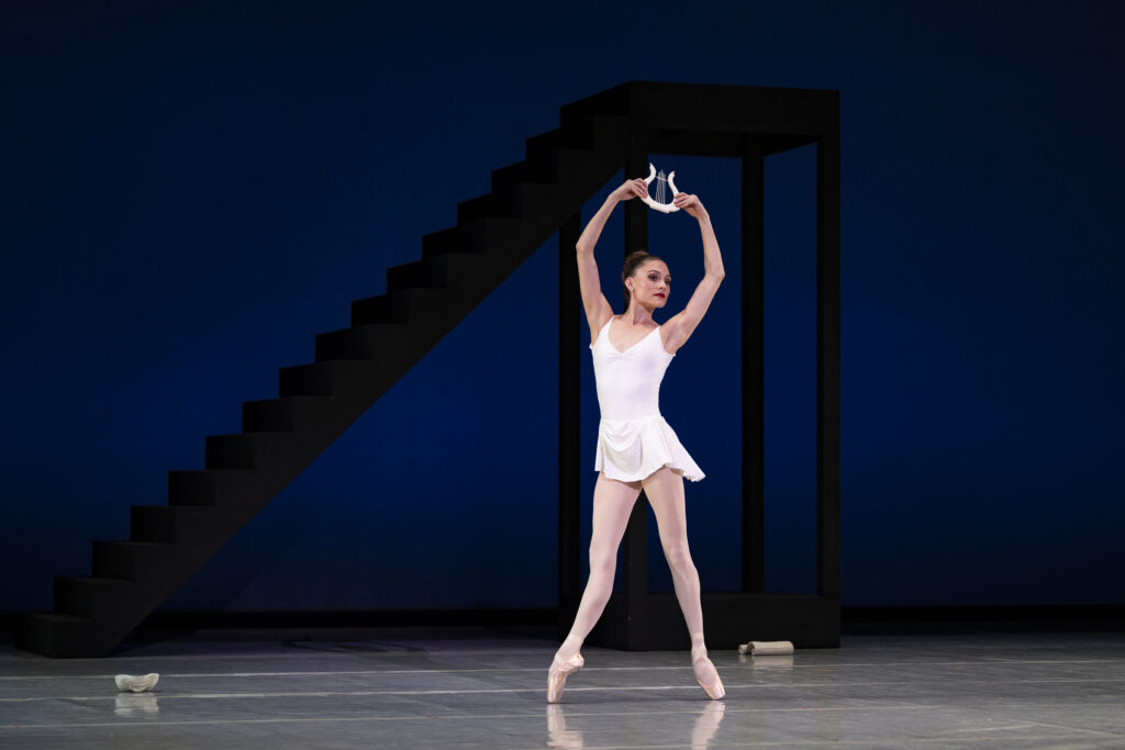 Amy Potter stands in fourth position effacé en pointe, arms raised above in a high fifth while holding a small lyre prop. She wears a white leotard and a short white skirt with pink tights. Behind her is a dark blue backdrop with a staircase set piece.