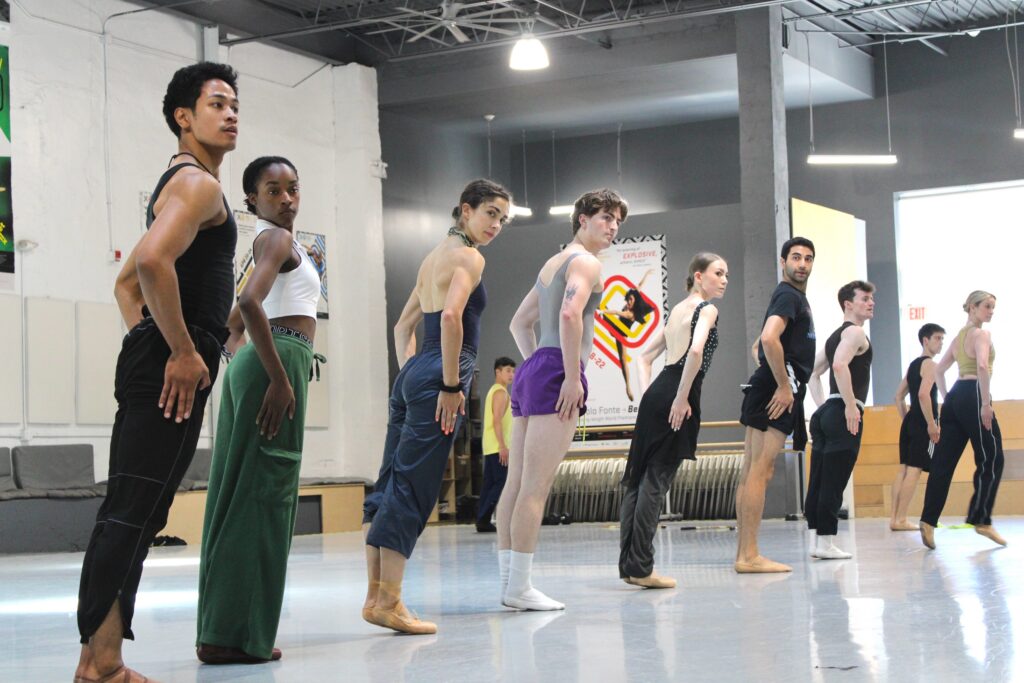 In a studio, nine dancers appear in a line, standing in parallel and leaning forward. Their arms are low and bent, with their hands placed on the outside of their upper thighs.