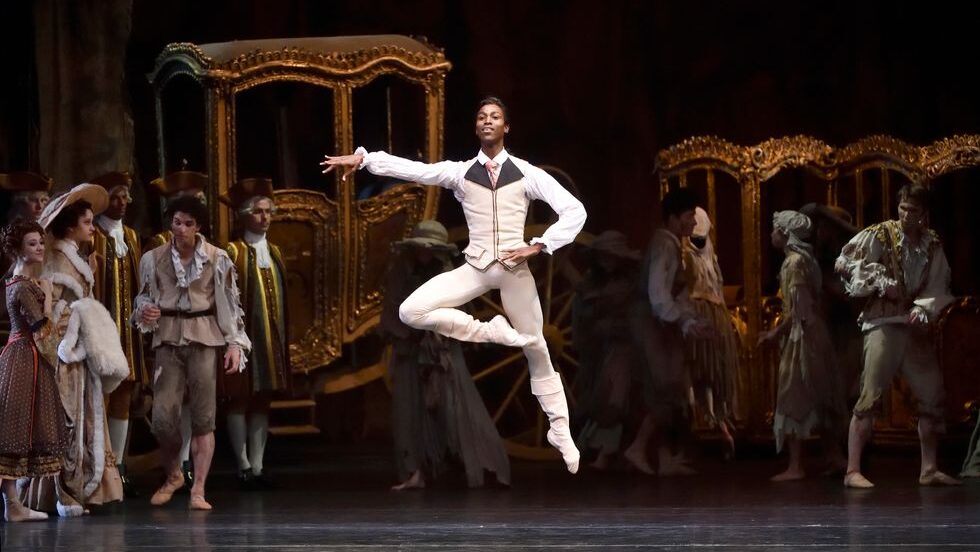 Calvin Royal III floats in the air with one leg in a high retiré devant as the other extends down and long. He rests one hand on his hip and reaches the other to the side. He wears white tights and boots, a white long-sleeve shirt, and an off-white vest with gold seams and a dark brown collar. Behind him, corps dancers dressed in 18th-century Parisian-style clothing, with gilded carriages among them.