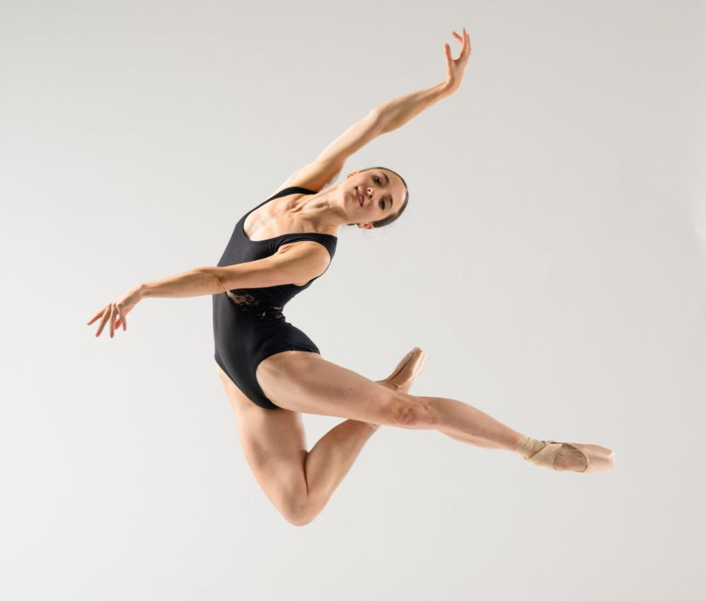 Carly Wheaton jumps with one leg tucked under and the other extended long in arabesque. She arches back while reaching one arm front and one above, smiling gently at the camera. She wears a black camisole leotard with a lace cutout on the sides.