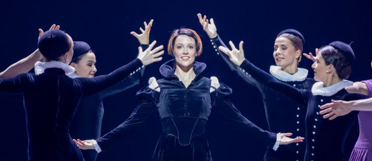 A woman with a short, dark bob with bright red streaks stands and smiles, her arms in demi second with her palms facing up. She wears a black velvet dress with long, puffed sleeves and a high collar. Four women, also in black velvet costumes and hats, surround her with their arms up.