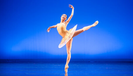 Onstage in front of a bright blue backdrop, Aina Oki does a piqué attitude derriere on pointe, her arms in a high third. She wears a bright gold pancake tutu.