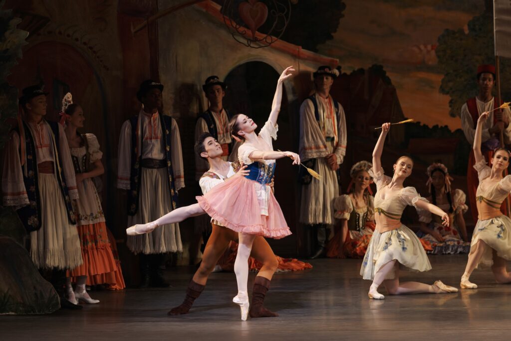 Megan Fairchild and Anthony Huxley perform a partnered arabesque onstage during a crowded town scene in the ballet Coppelia. Fairchild wears a knee-length dress with a pink skirt, blue bodice, and puffy white sleeves as she balances in third arabesque on pointe. Huxley wears russet tights and brown ballet boots and a white peasant blouse and he holds her by the waist. Other dancers pose in the background.