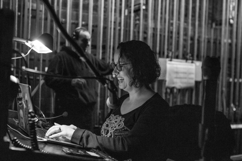 In this black and white photo, Courtney DiBello sits at a podium backstage at a theater and speaks into a microphone as she types on a laptop keyboard.