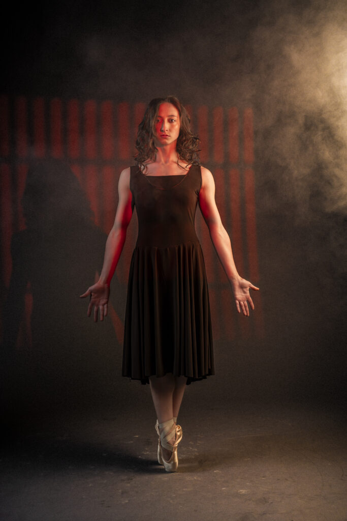Alexandra Troianos stands in sous-sus en pointe with her arms extended long, palms flat and forward, as she stares at the camera with a blank expression. She wears a long dark brown dress and keeps her curly brown hair down. Behind her is a jail cell with haze.