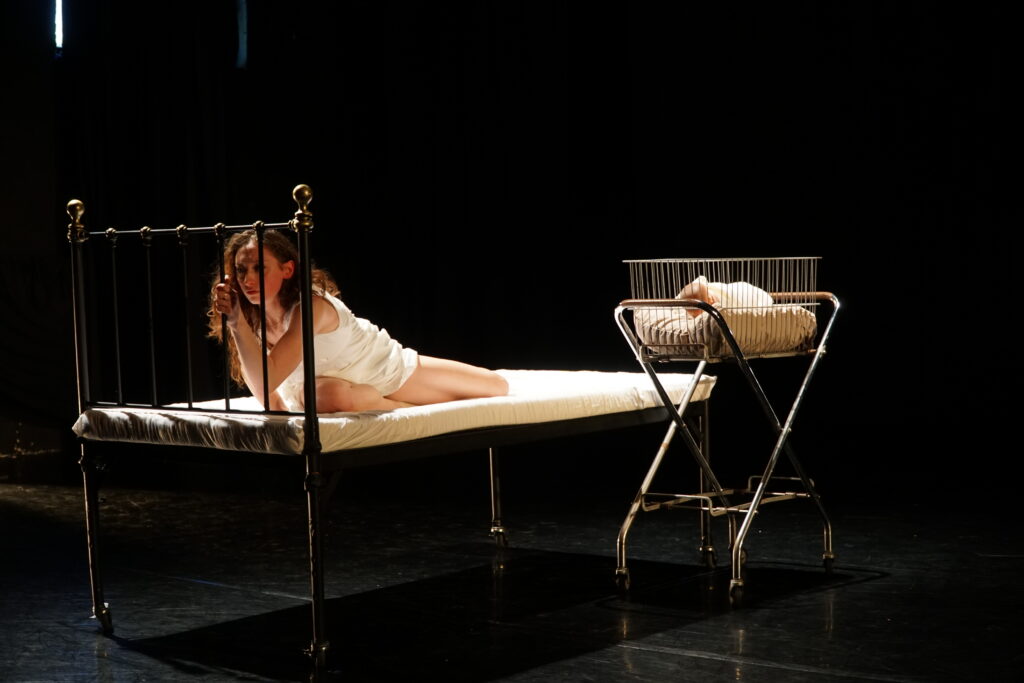 On a dark stage, Alexandra Troianos sits on a bare hospital bed as she twists her body toward the metal headboard, clasping one metal rod with her hand. She looks depressed and trapped. Beside the bed, in a cradle, is an infant. 