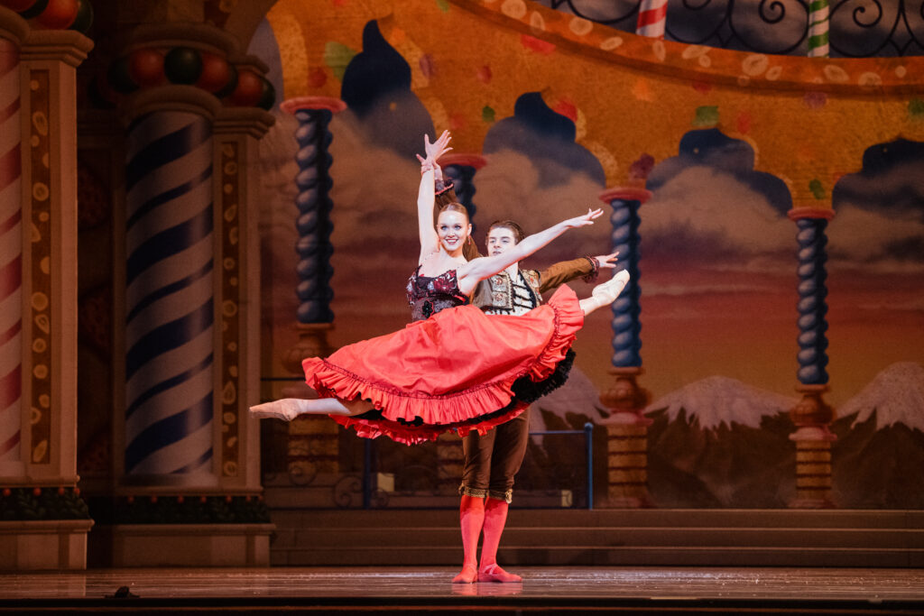Two dancers perform the "Spanish" divertissement from "The Nutcracker." Adriana Wagenveld, wearing a long red dress with a full skirt, splits her legs with her arms lifted as her partner, standing behind her, holds the wrist of her top arm gently.