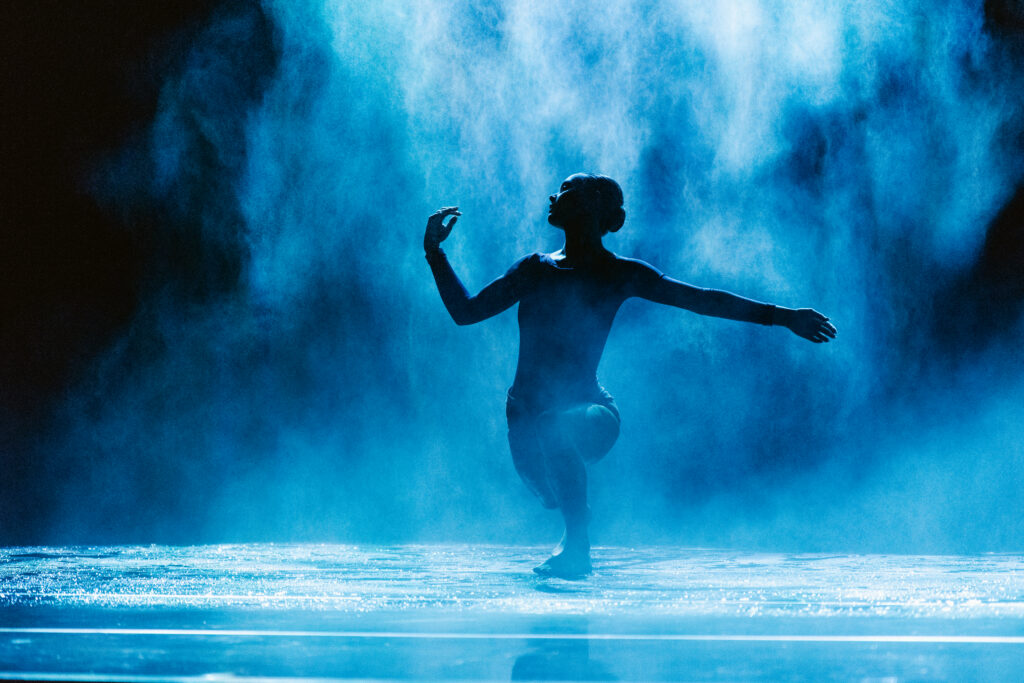 Obscured partially by a haze of mist, Adriana Wagenveld lunges forward while turning her head in profile, one arm extended to the side as the other bends at the elbow. She is visible in silhouette as stage lighting illuminates a cloud of mist behind her and the slick floor.