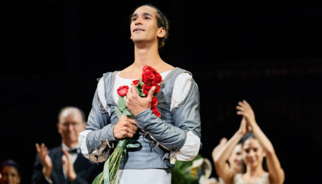 Davi Ramos, dressed in light gray tights and a gray and white tunic, clutches a bouquet of roses to his chest as he looks out toward the audience with emotion. Behind him, dancers and artistic leadership clap to celebrate his promotion.