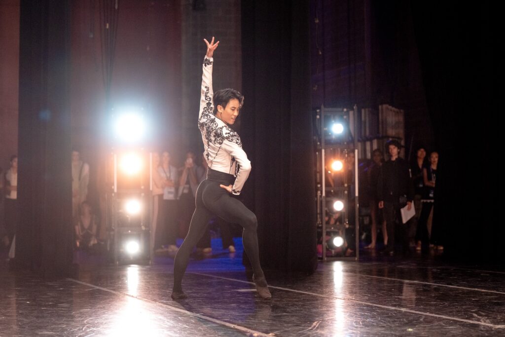 A young male ballet dancer wearing a white bolero jacket, black tights, and black ballet shoes poses confidently onstage during a performance. He faces upstage in a wide fourth, his right foot pushed onto demi pointe, with his right hand on his waist and his left arm up.