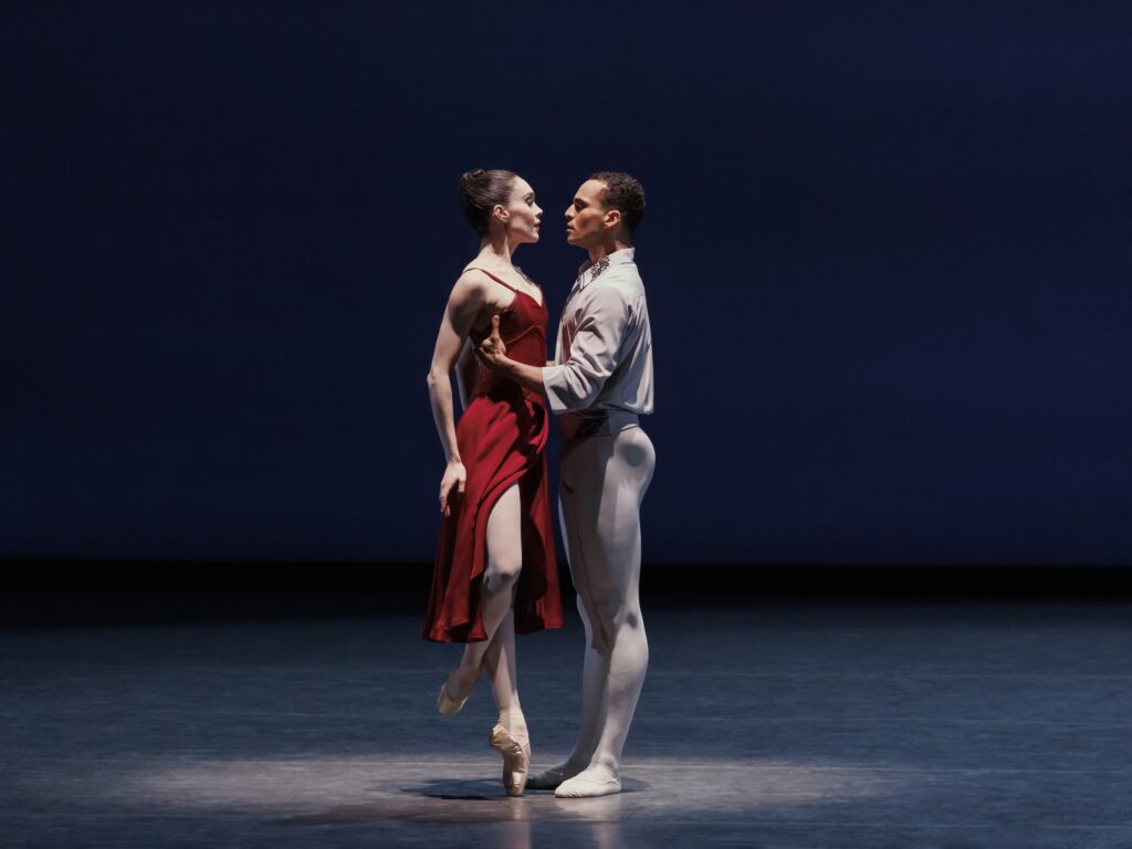 A couple performs a pas de deux. The woman, wearing a calf-length red dress, faces the man and stands on pointe in coupé derriére, her arms down by her sides. Her partner, wearing a white shirt and tights, holds closely her by the waist. They look at each other intensely.