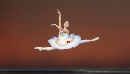Isabella Padilla does a saut de chat onstage with her arms in high third. She wears a white pancake tutu with gold ornamentation and two red roses at her hip, with a third rose in her hair.