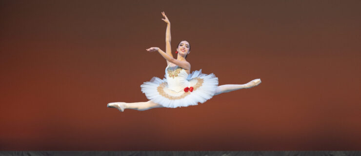Isabella Padilla does a saut de chat onstage with her arms in high third. She wears a white pancake tutu with gold ornamentation and two red roses at her hip, with a third rose in her hair.