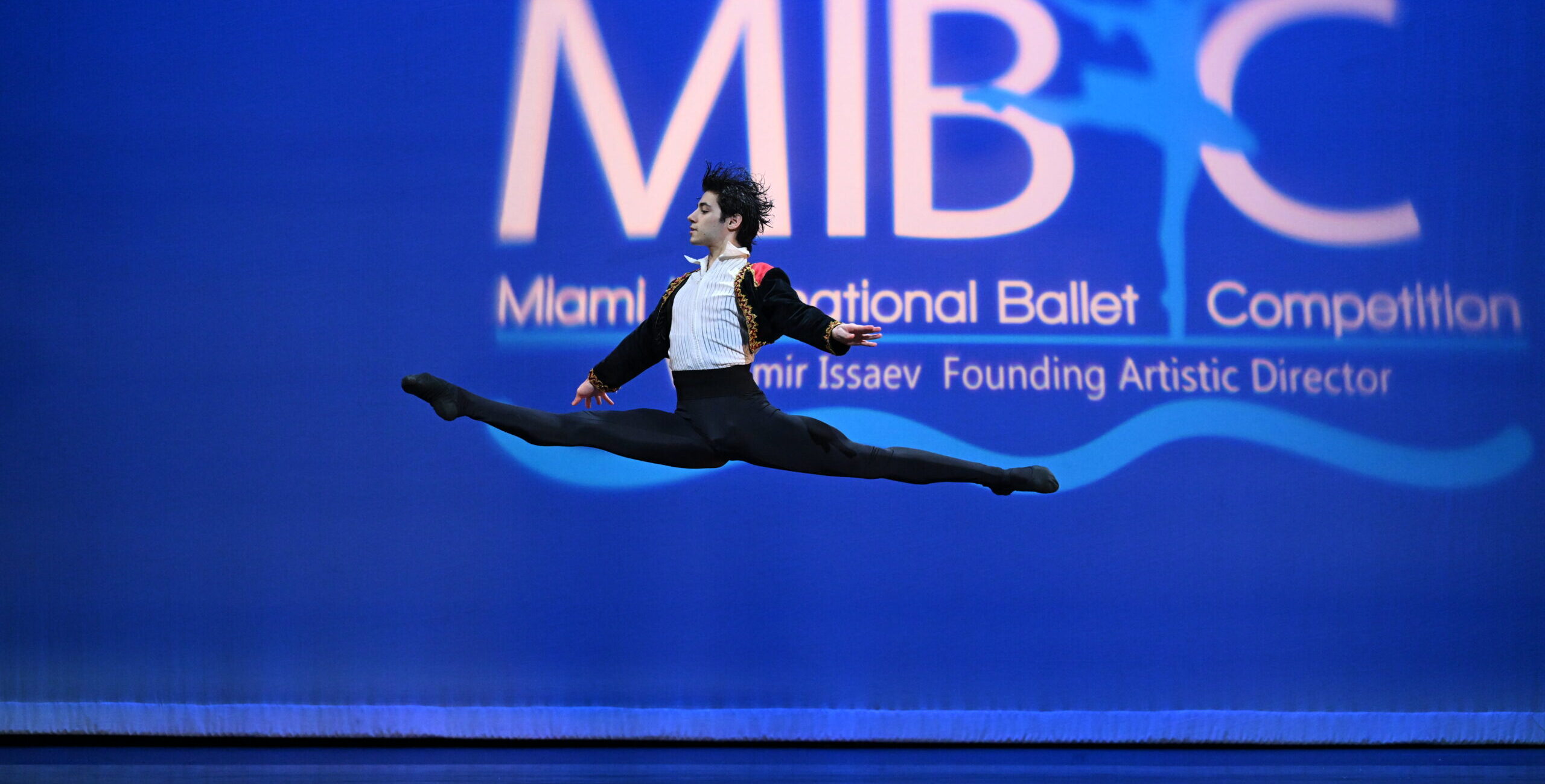 Pablo Acosta splits his legs mid-air with his arms low to either side. He wears black tights, a white button-up, and a black tunic with red and gold details.