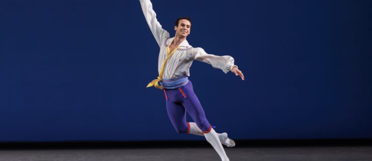 Ryan Tomash does a jeté with his arms extended in a diagonal line. He wears a billowy white blouse, light blue cummerbund, blue knee-length tights over white tights, and white ballet slippers. He performs onstage in front of a blue backdrop.